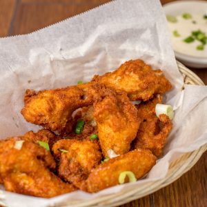Buffalo Chicken Wings in a woven basket, sitting in wax paper with a side of green onion sprinkled ranch in a ceramic bowl on a wooden table.